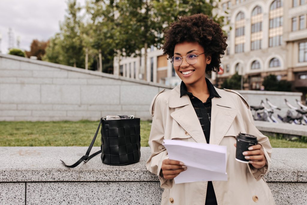 attractive dark skinned woman beige trench coat eyeglasses smiles holds coffee cup paper sheet outdoors